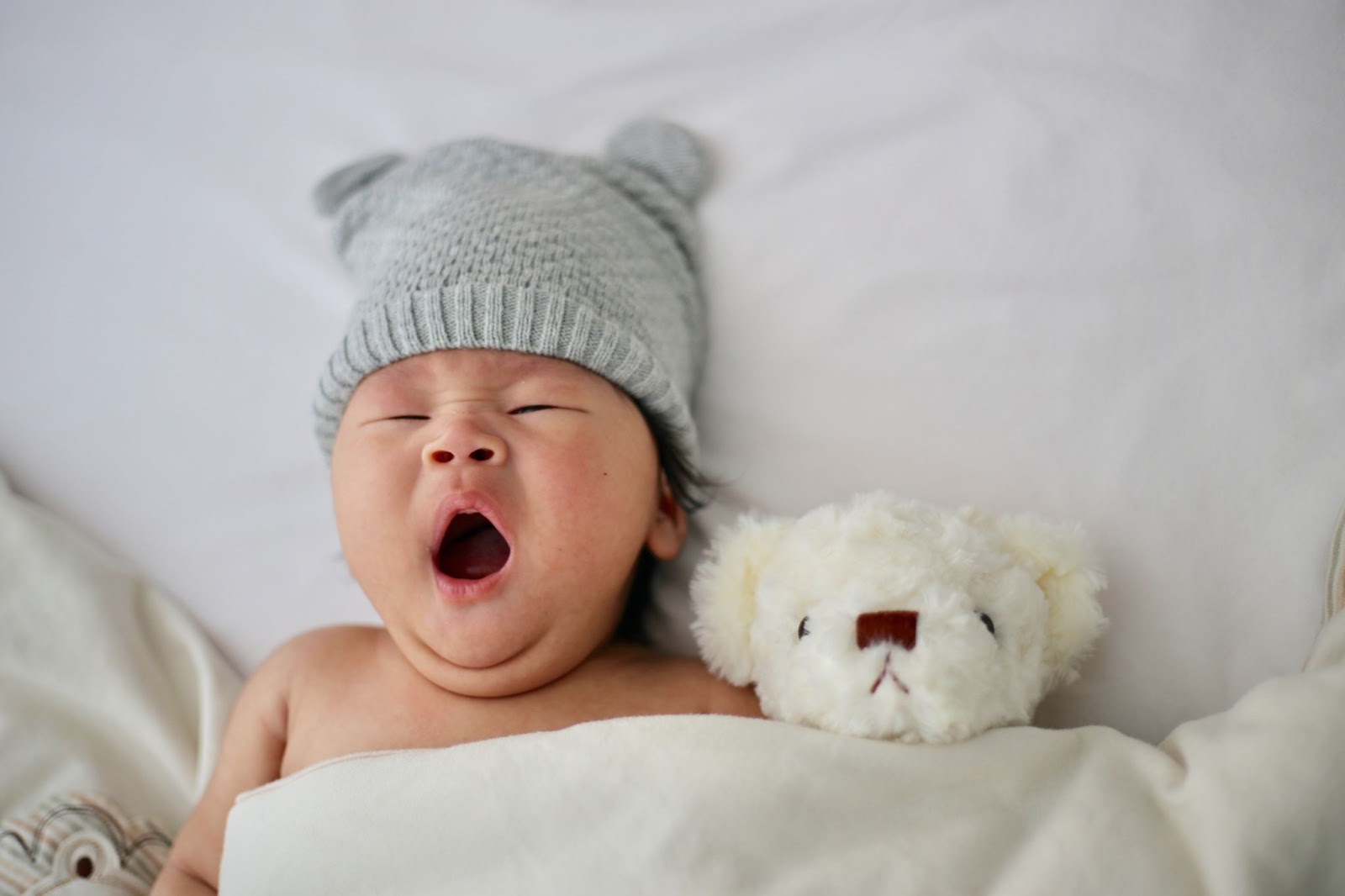 Un bebé joven bostezando en una cama con un osito de peluche.