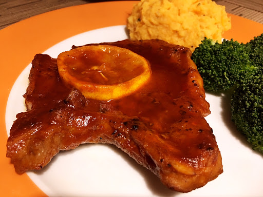 A pork chop on a plate topped with sauce and a slice of lemon with broccoli and mashed potatoes.