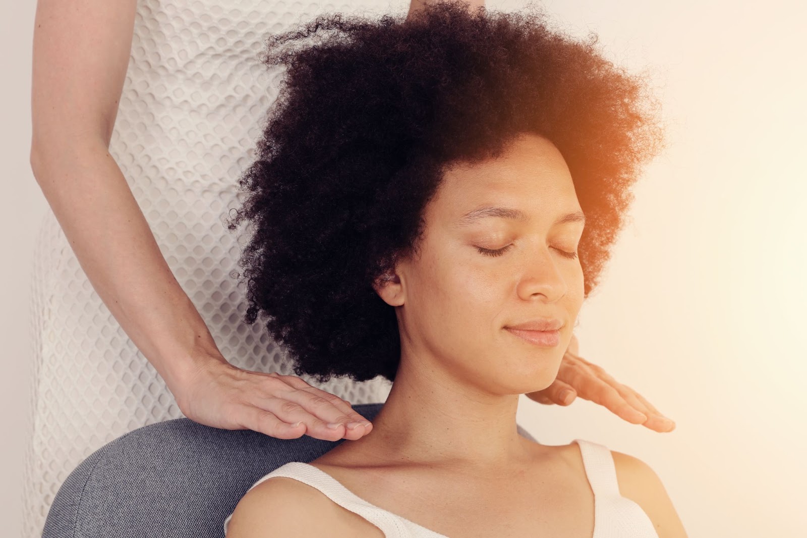 A woman sitting upright receiving Reiki treatment as a woman holds her hands above her shoulders. 