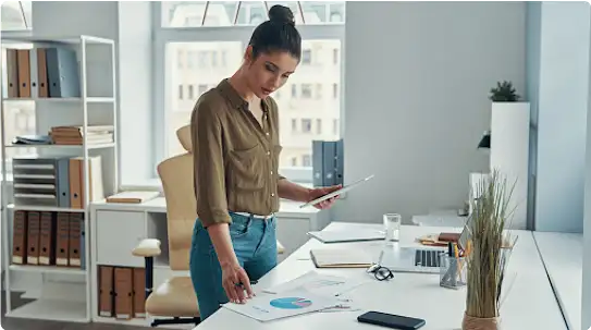 A young woman stands at a desk, with a tablet in her left hand, looking down at a printed pie chart to her right.