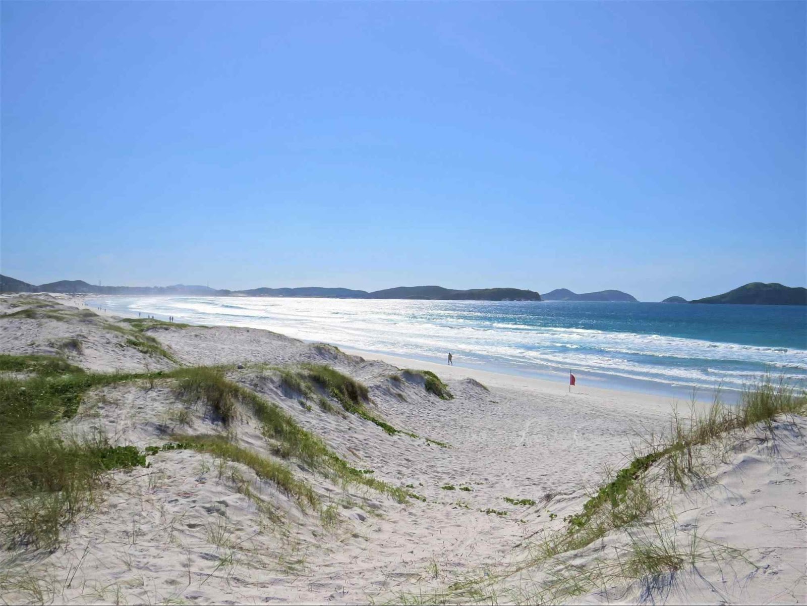 Grandes dunas de areia em frente ao mar na Praia das Dunas. Pequenos rastros de vegetação no topo das areias brancas. Céu azul sem nuvens, mar azul com ondas e montanhas ao fundo da paisagem.