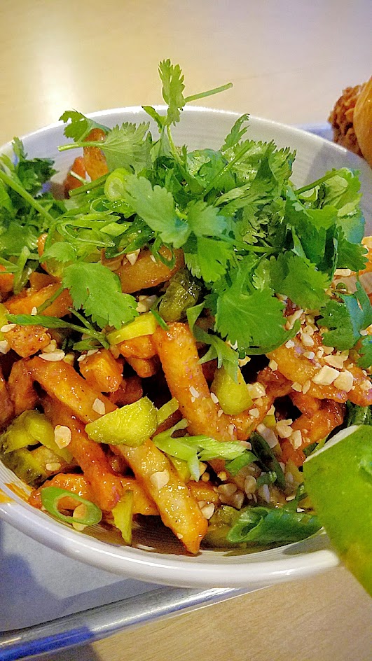 Basilisk vegan Dan Dan fries, Dan Dan fries, a large bowl of hand cut fries covered in peanut sauce, chili oil, baby dill pickles, crushed peanuts, green onions, cilantro and lime