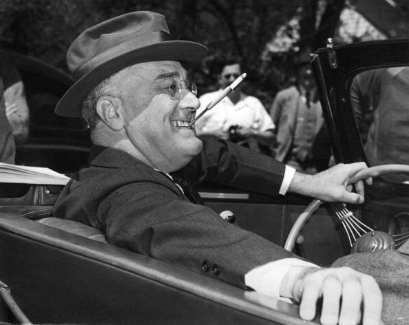 With a cigarette in a holder clenched in his teeth a smiling Franklin Delano Roosevelt sits jauntily at the wheel of his convertible Warm Springs...