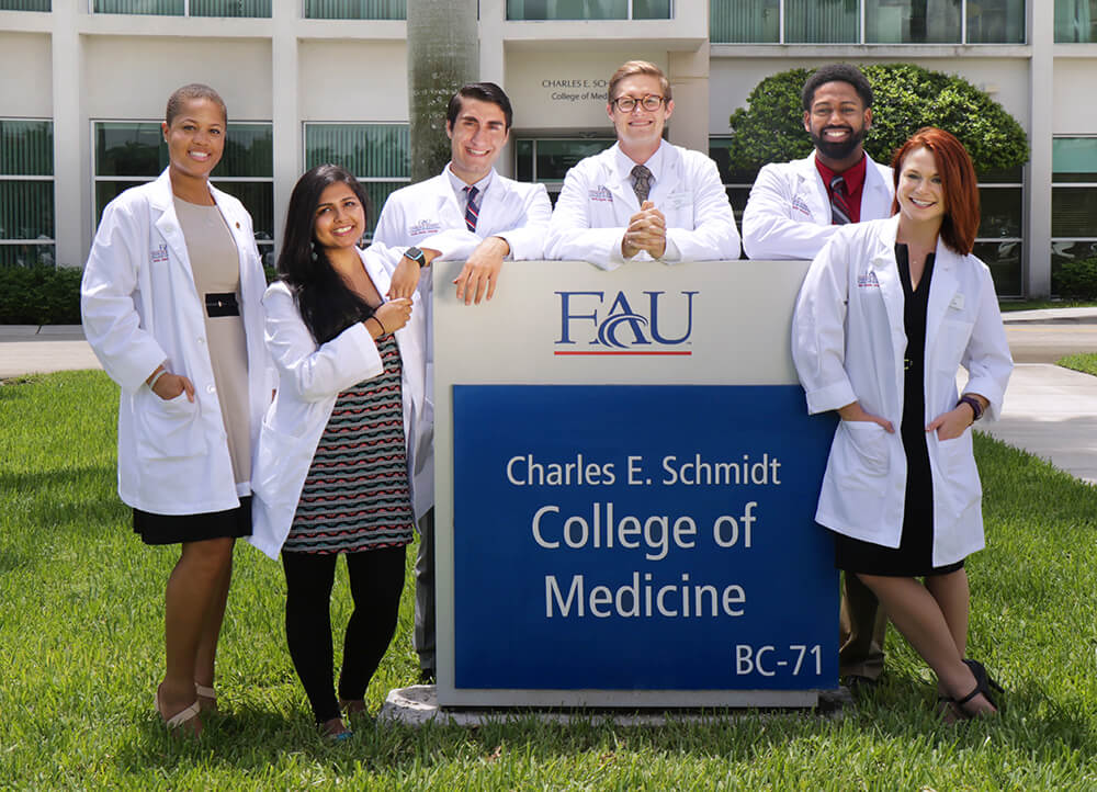 A group of medical students are standing outside the Charles E. Schmidt College of Medicine building, posing around a sign. They are all wearing white coats and appear to be discussing their studies.