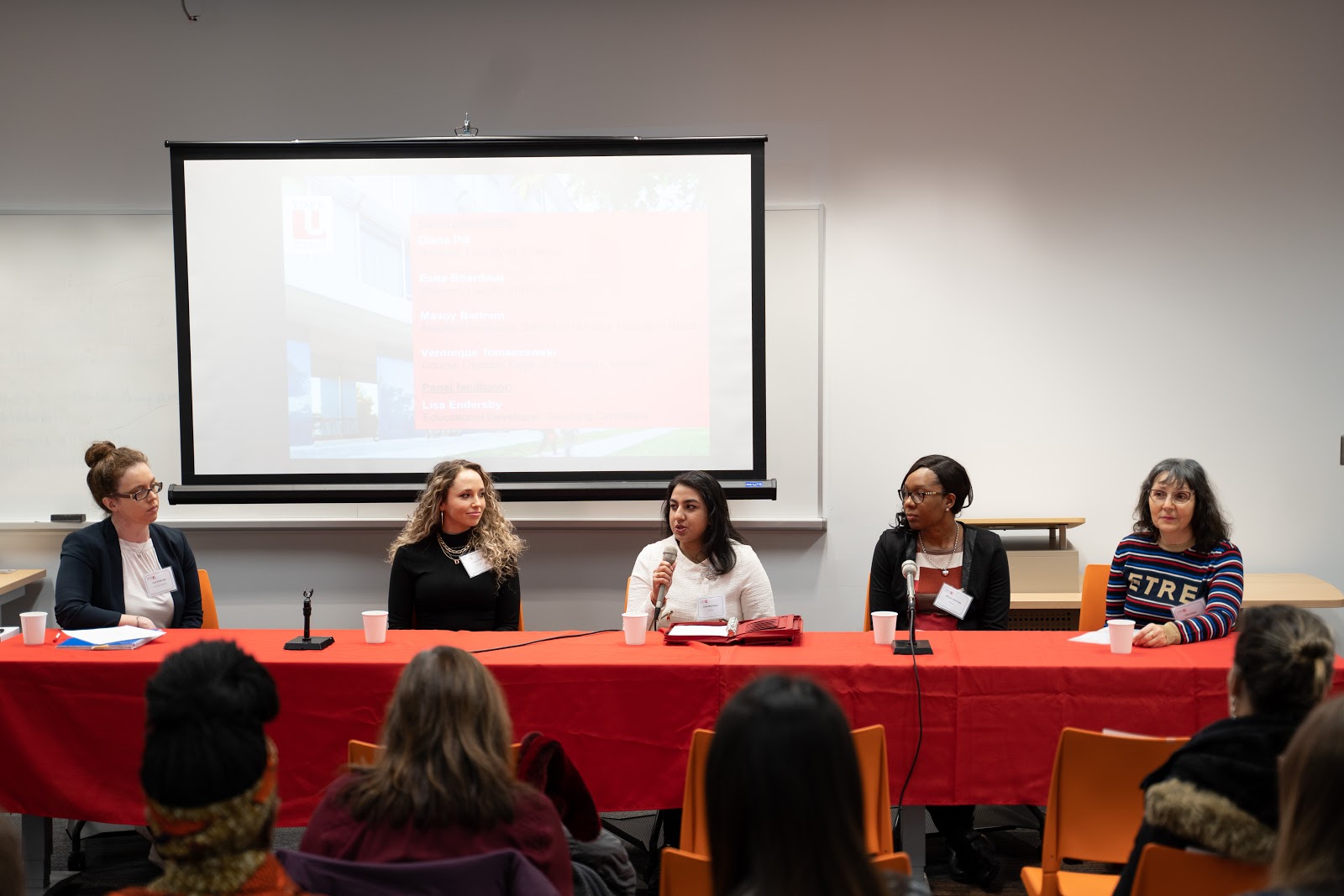 5 people sitting at a table, four of which are panelists. The individual in the middle is holding a microphone. 