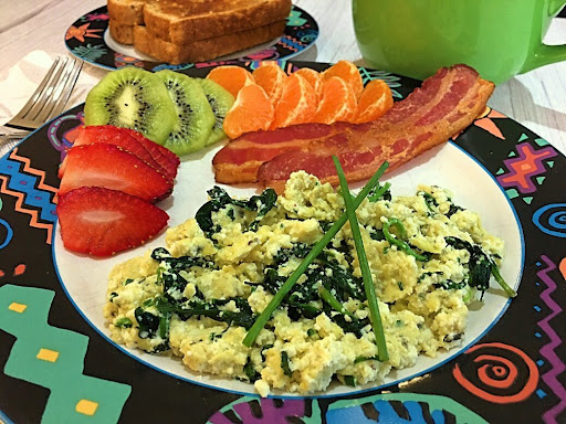 A gorgeous plate of Spinach & Ricotta Scrambled Eggs along with crispy bacon and fruits.