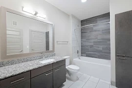 Modern apartment bathroom with tile floors, a long vanity next to the toilet, and a tub/shower combo with gray tile surround.