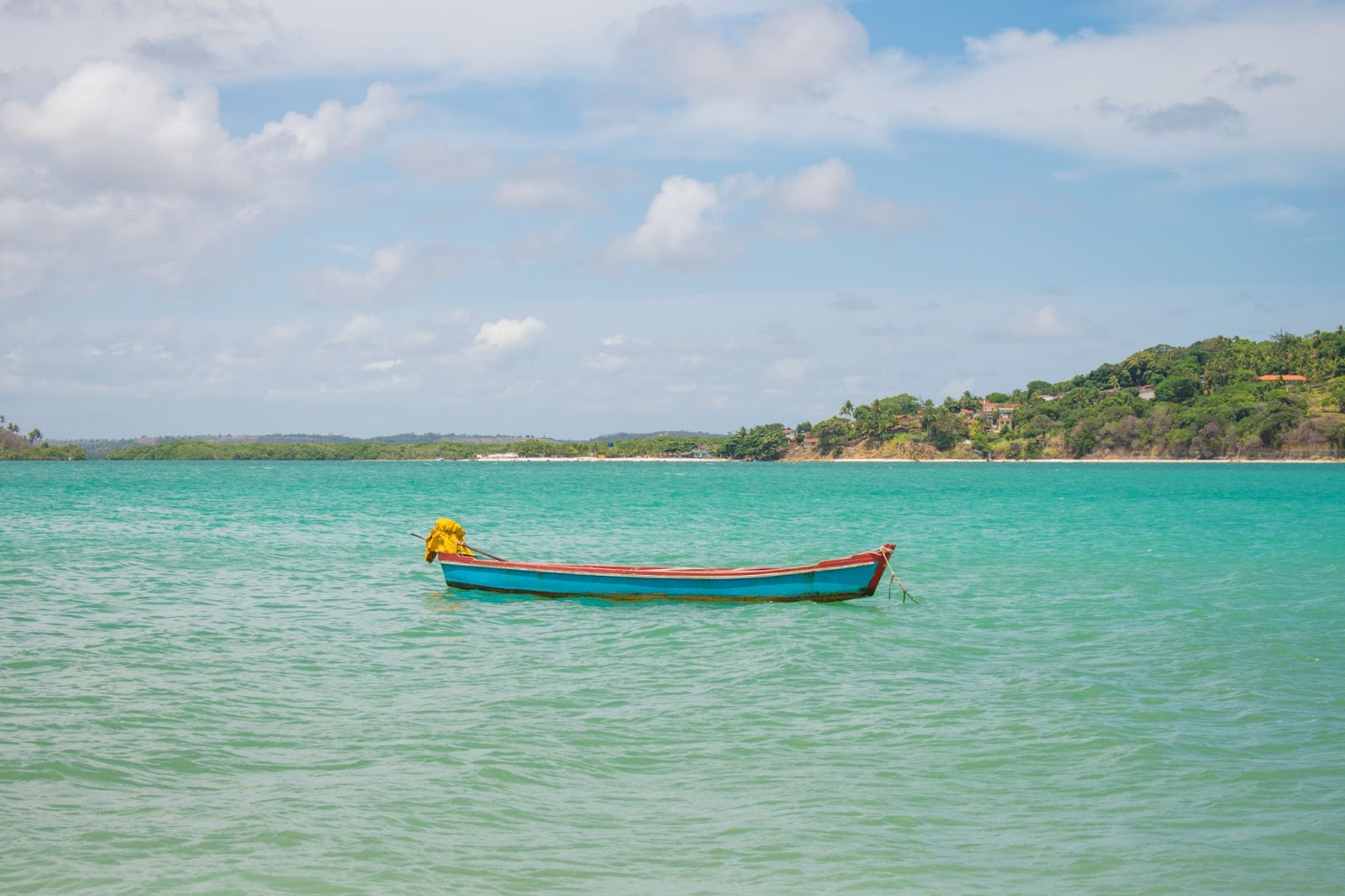 Barco em mar azul e sem ondas. Faixa de areia ao fundo com pequena montanha com vegetação verde.