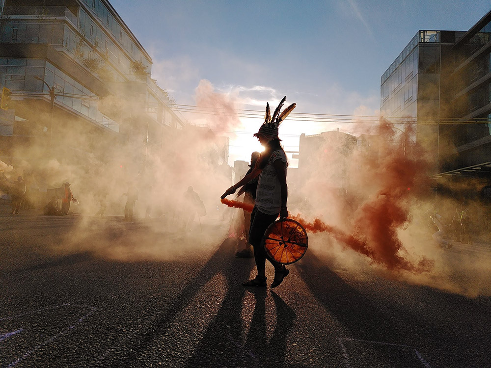 A rebel in feather hat and holding a smoke flare that clouds the scene in orange is shillouetted by the sun.