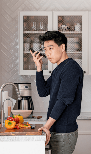 An Asian American man stands at a kitchen counter holding his Android smart phone up to his ear.