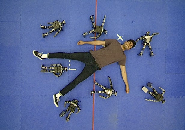 A photo of Akhil Raju lying on a blue floor. He is surrounded by small models of robots.