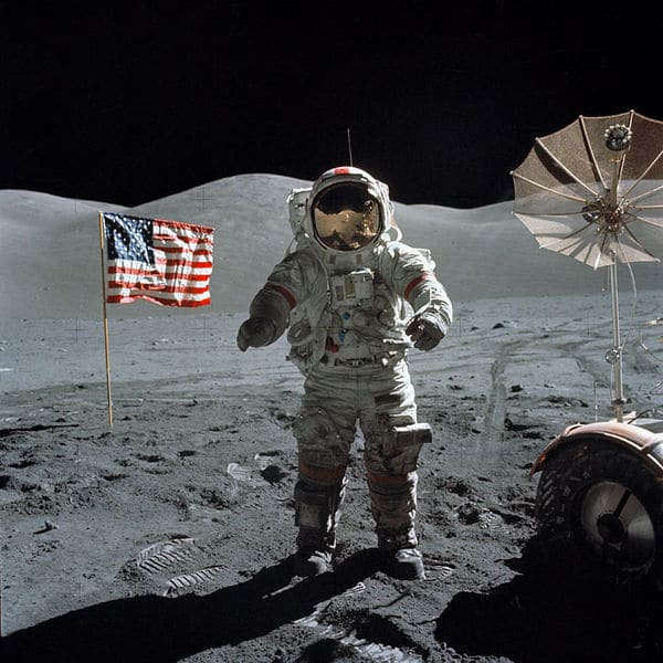 A Astronaut standing on a moon with American flag and flash on back.