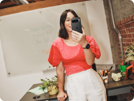 Woman in a bright pink top is holding a pixel 9 in her hand while leaning on her desk.