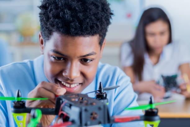 Cheerful high school student concentrates while building a drone Happy African American high school student smiles cheerfully while building a drone during engineering class. A female student is working in the background. students and drones stock pictures, royalty-free photos & images