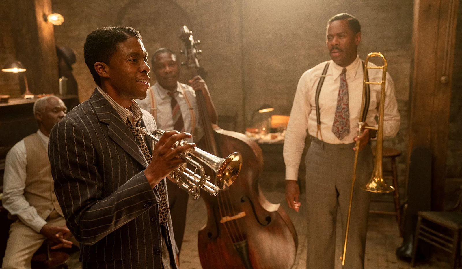 This is a screen still from Ma Raineys Black Bottom. Chadwick Boseman stands in the foreground, laughing and holding a trumpet.