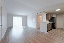 Spacious, empty apartment interior with wood-style flooring, white walls, and modern kitchen appliances.