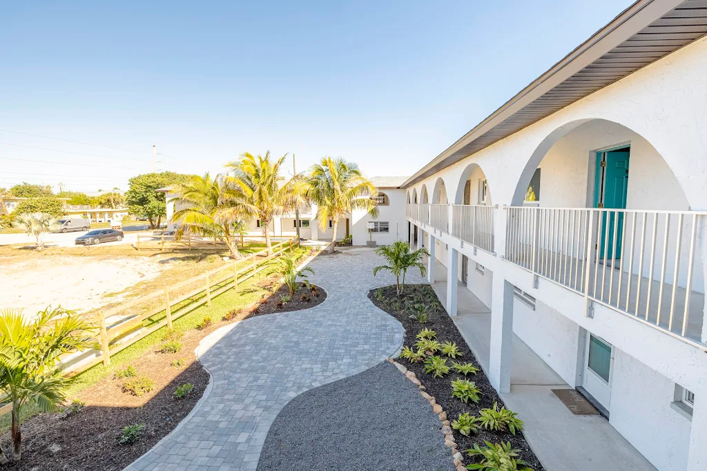 Two-story apartment complex with arched doorways, white railings, a curved pathway, and landscaped garden in daylight.