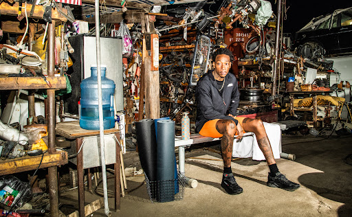 Wiz Khalifa sitting on a bench in a car scrapyard.