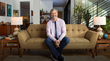 American director, writer, and producer Judd Apatow wearing a pink and blue plaid shirt and blue t shirt, sitting on a couch, smiling to the camera