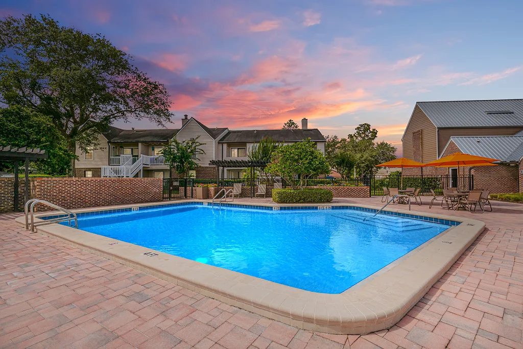 Swimming pool with picnic tables, lush landscaping, and apartment buildings in the background at dusk