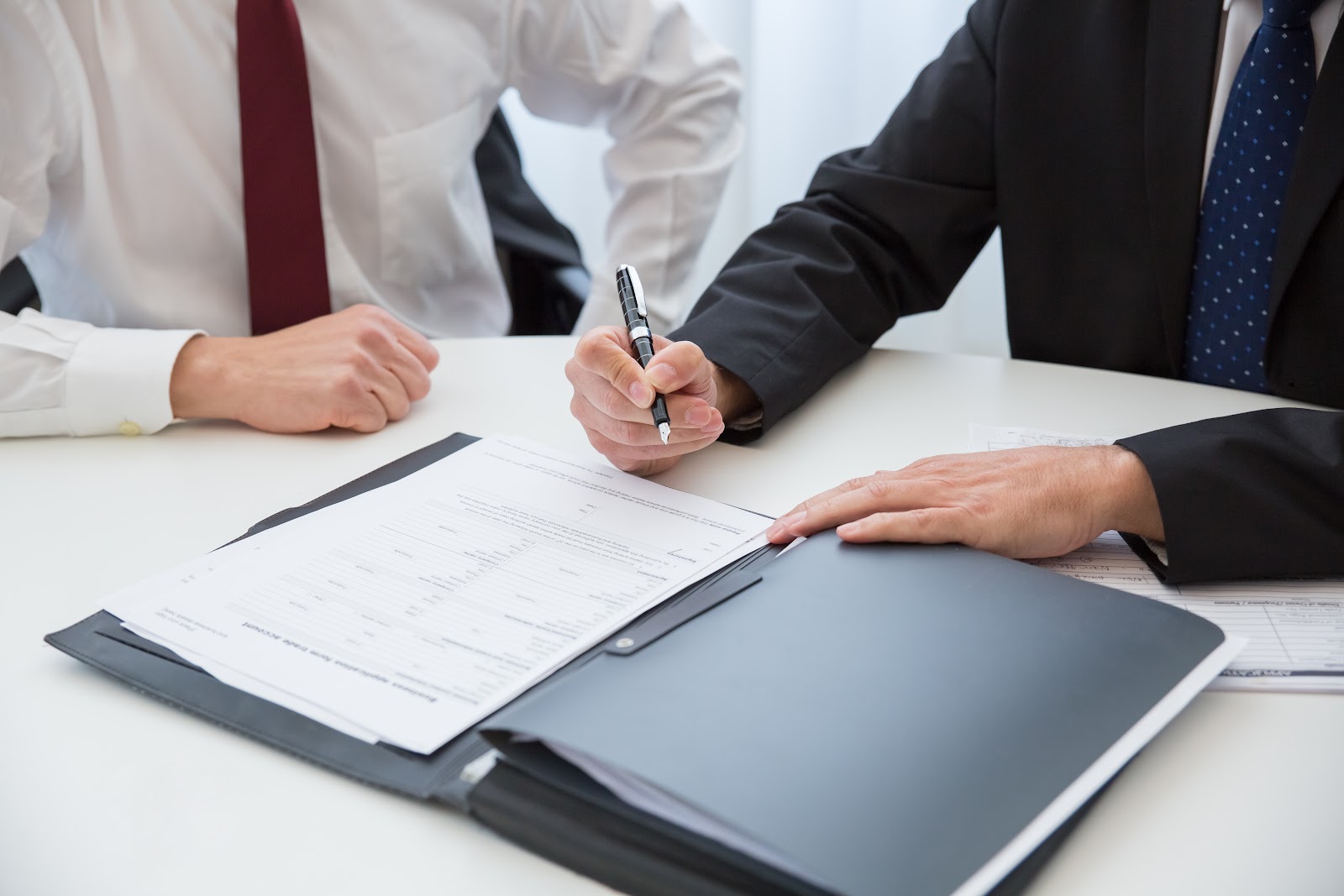 A Person in Black Suit Holding a Pen Near the Documents on the Table