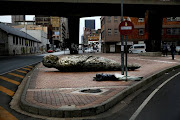 A man sleeps out on the street, on a gloomy day, next to a piece entitled Maboneng Man, by local artist, Ledelle Moe, this forms part of her Displacements series. Maboneng, a formerly poorer and less developed area in inner city Johannesburg has been undergoing a process of gentrification.