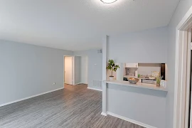 Livingroom with grey wood flooring & white walls, small opening over bar area furnished with plants looks into kitchen.