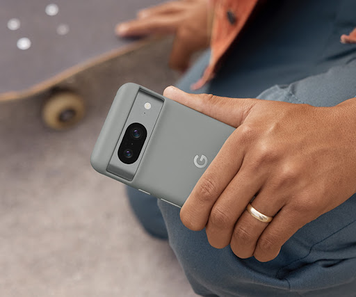 A person kneels beside their skateboard. In their left hand is a Pixel 8 in a Pixel 8 Case. They also wear a Pixel smartwatch.