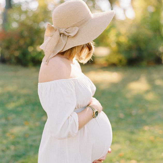 A fair skinned woman with short blonde hair stands in the grass, looking away from the camera, with her arms hugging her pregnant belly. She wears a white dress and a tan sun hat.