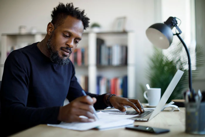 A man sitting at his work desk, using a laptop and taking notes on the side.