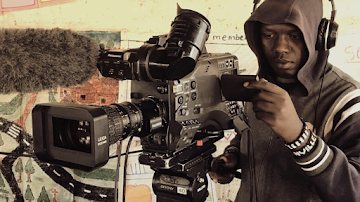 A young man stands behind a large video camera with multiple viewports and a boom microphone.