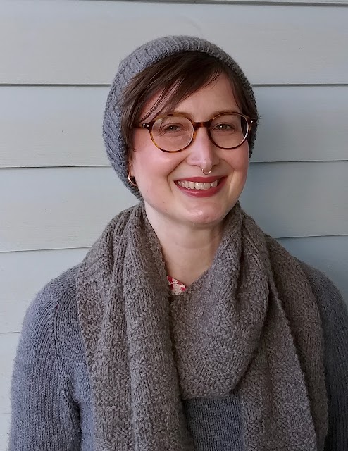 Siobhan stands in front of a weatherboard wall, wearing a grey handknit textured hat, similar style scarf, and plain grey handknit jumper. She is smiling.