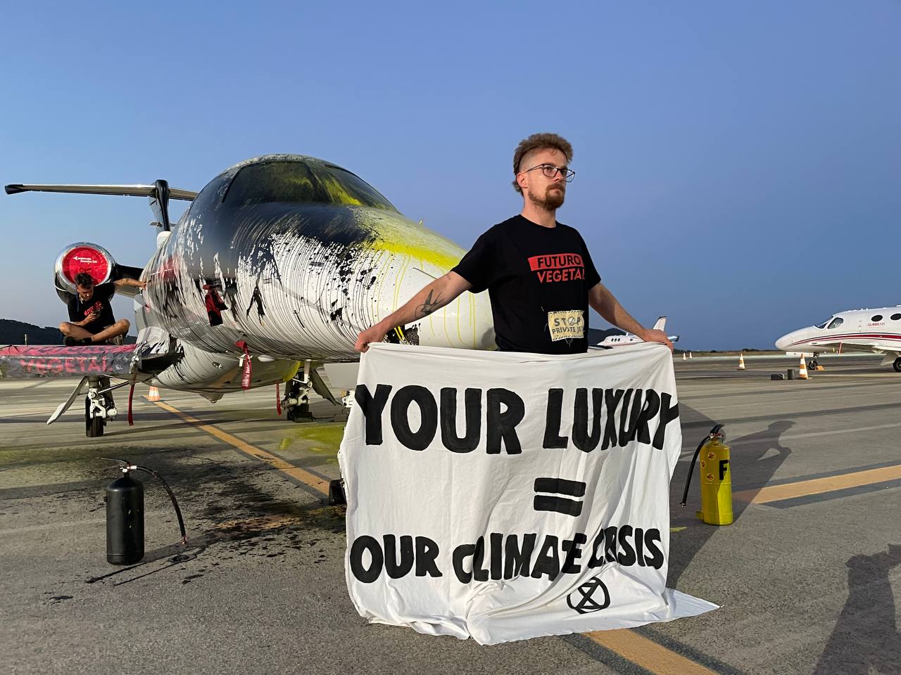 An activist holds a banner (Your Luxury = our climate crisis) beside a private jet covered in black and yellow paint