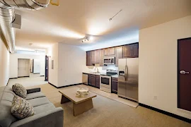 Furnished living and kitchen areas with exposed ductwork, dark wood cabinets, stainless steel appliances, and white walls