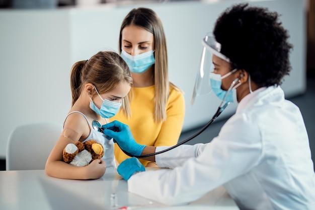 Pediatra examinando menina com estetoscópio durante pandemia de coronavírus em uma consulta de assistência médica ambulatorial