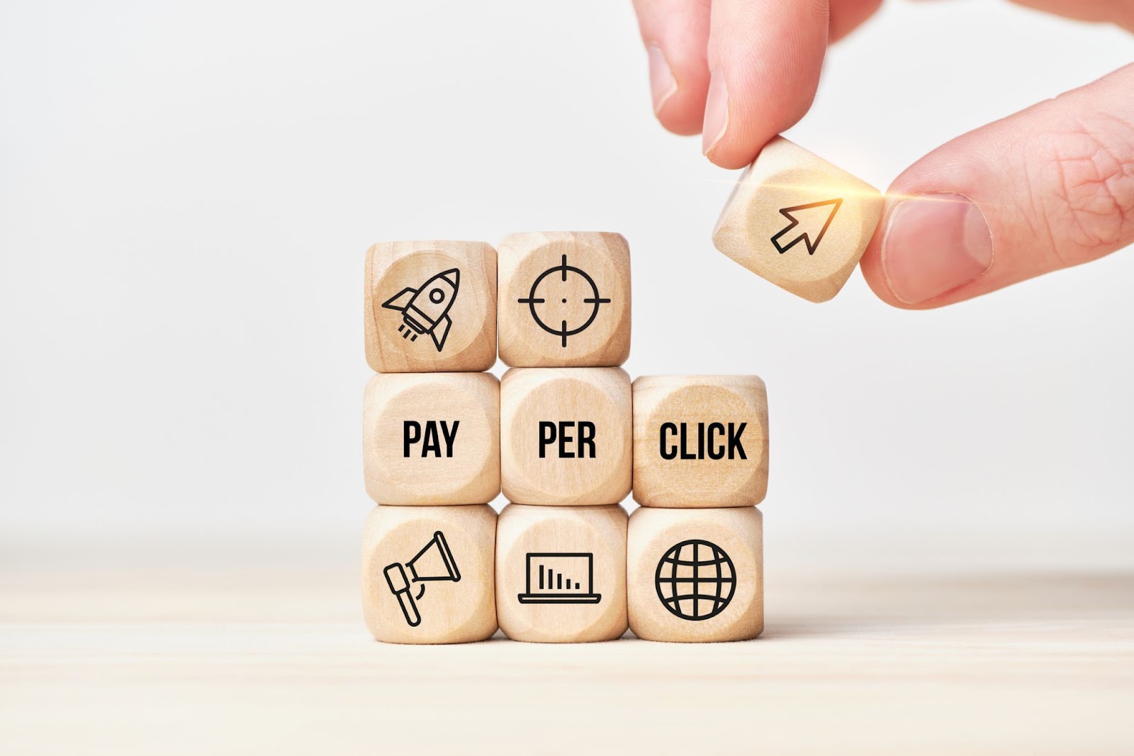 Fingers grabbing a wooden block with an icon on it from a pile of other blocks, each with various digital marketing icons, and in the center reads: “Pay-per-click”. 