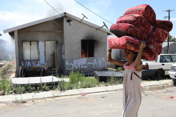 A young woman carrying sofa cushions walks past a burning RDP house after fighting at the Fynbos housing development.