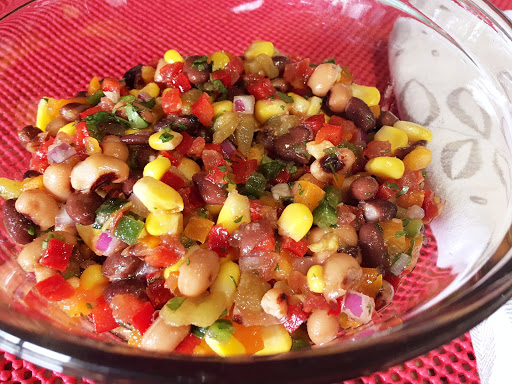 Beans with other vegetables in a glass serving bowl sitting on a red table mat next to a white napkin.