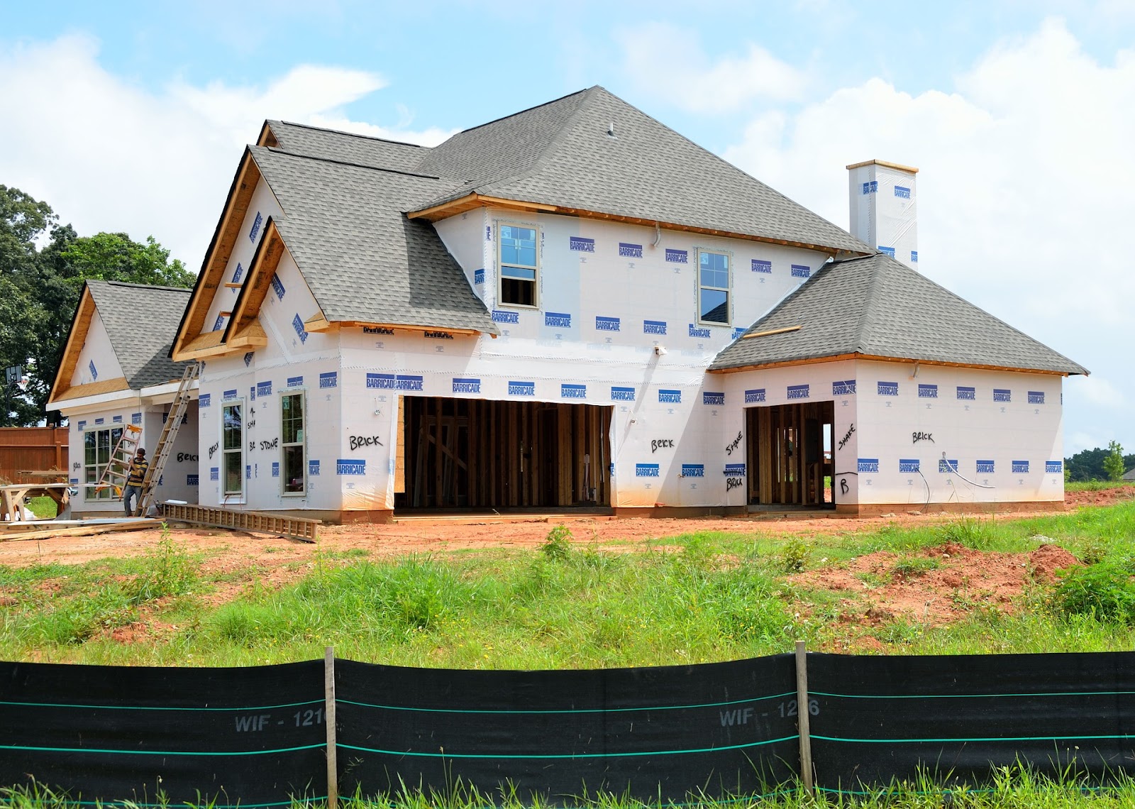 An entire home is being constructed.  The frame, walls and roof are complete.  House-wrap coats the outside and a construction worker leans a ladder against the front of the home.