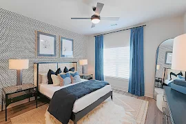 Model unit bedroom with wallpapered accent wall, large window with blinds and curtains, wood plank flooring, and modern decor