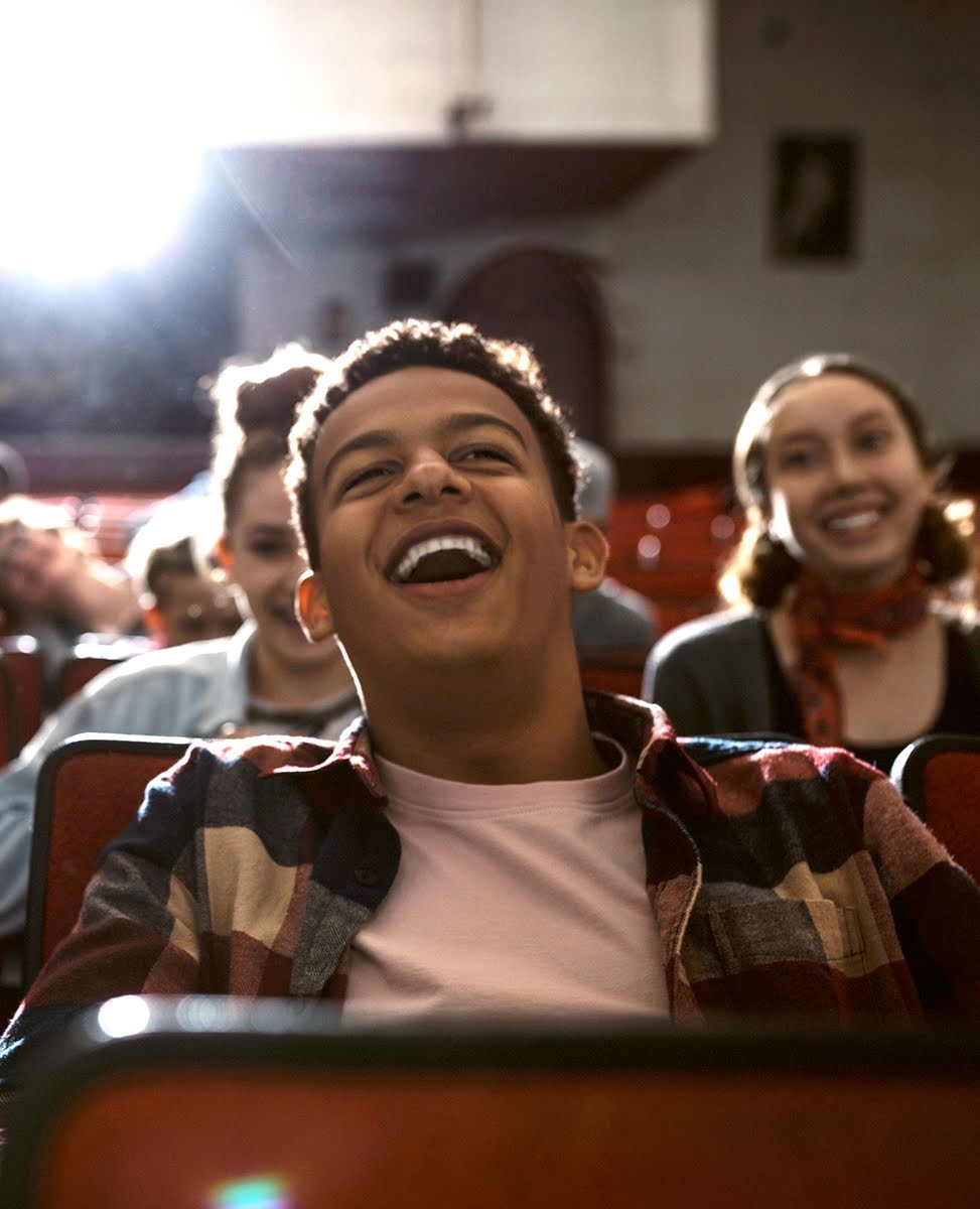 A teenage boy laughs among friends while sitting in a cinema.