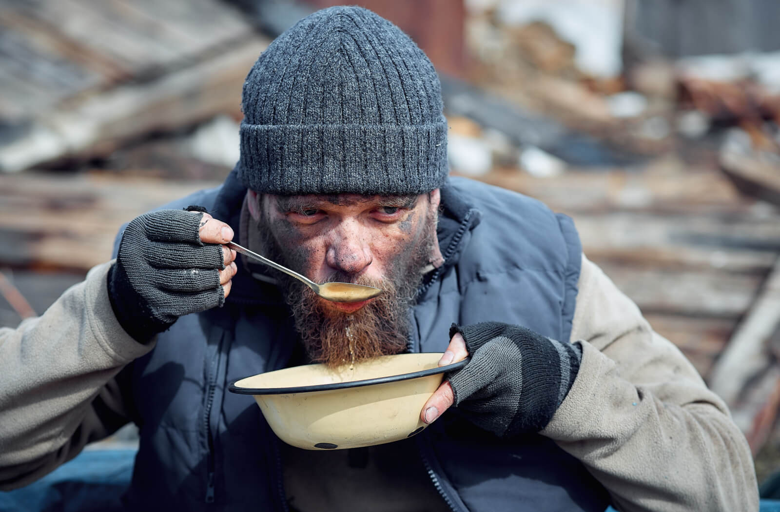 A homeless person wearing layers of clothes enjoying a bowl of soup.