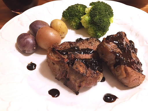 Two lamb chops with dark sauce drizzled on top on a white plate with baby potatoes and broccoli.