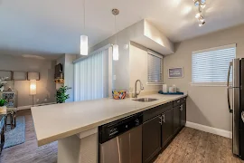 Modern apartment kitchen with stainless steel appliances, pendant lighting, windows with blinds, and a breakfast bar.