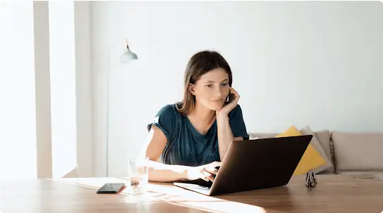 Uma mulher com uma blusa azul-escuro usa um laptop em uma mesa de madeira. Também estão sobre a mesa um smartphone, um copo de água e um par de óculos.