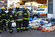 Firefighters stand next to covered bodies of victims at the scene of a deadly blaze in Johannesburg on August 31 2023.