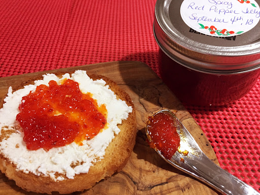 A cracker topped with cheese and red pepper jelly sitting on a wood cutting board along with a spoon and a mason jar in the background.