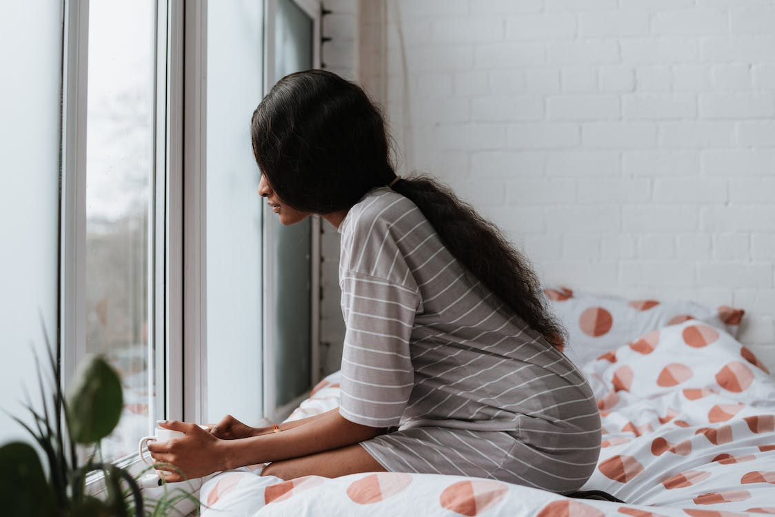 A Woman in Striped Shirt Sitting on the Bed Near the Glass Window Holding a Mug