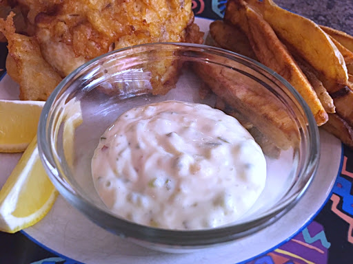 Sauce in a small glass serving bowl with lemon wedges, french fries and battered fish on a plate.