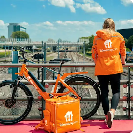 A Just Eat Takeaway delivery person stands on an overpass dressed in a branded orange windbreaker